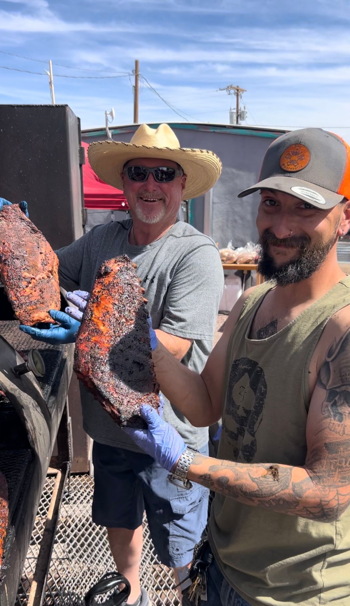 Two men outdoors handling a large cooked fish on a grill, smiling in hats and sunglasses.