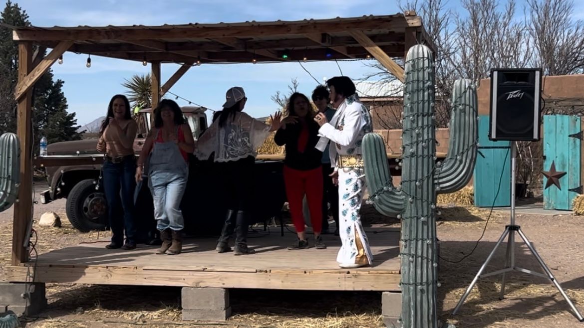 People dancing on a wooden stage under a rustic shelter in a desert setting.