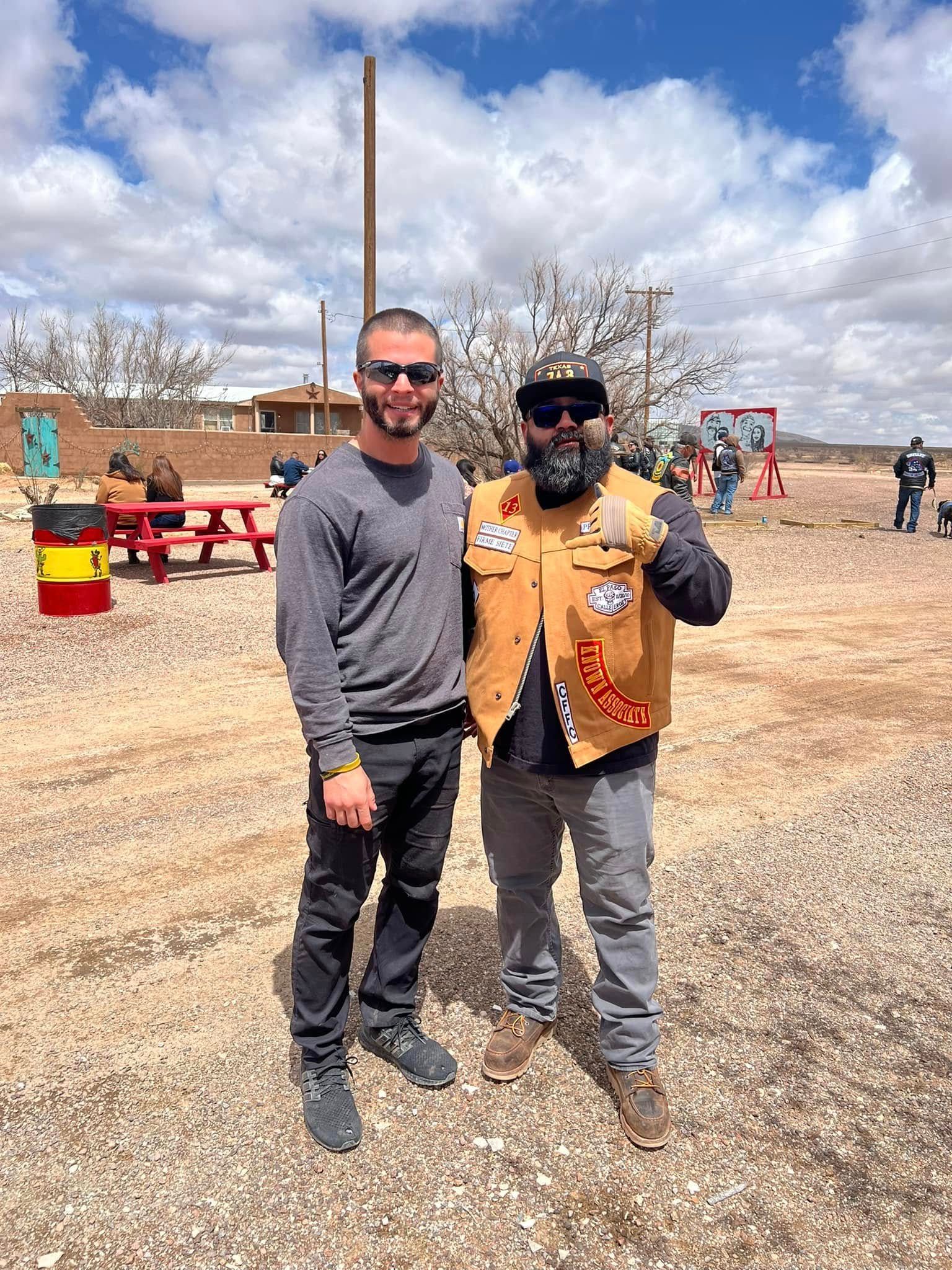 Two men standing together outdoors in a dusty field, one giving a thumbs-up and wearing a vest.