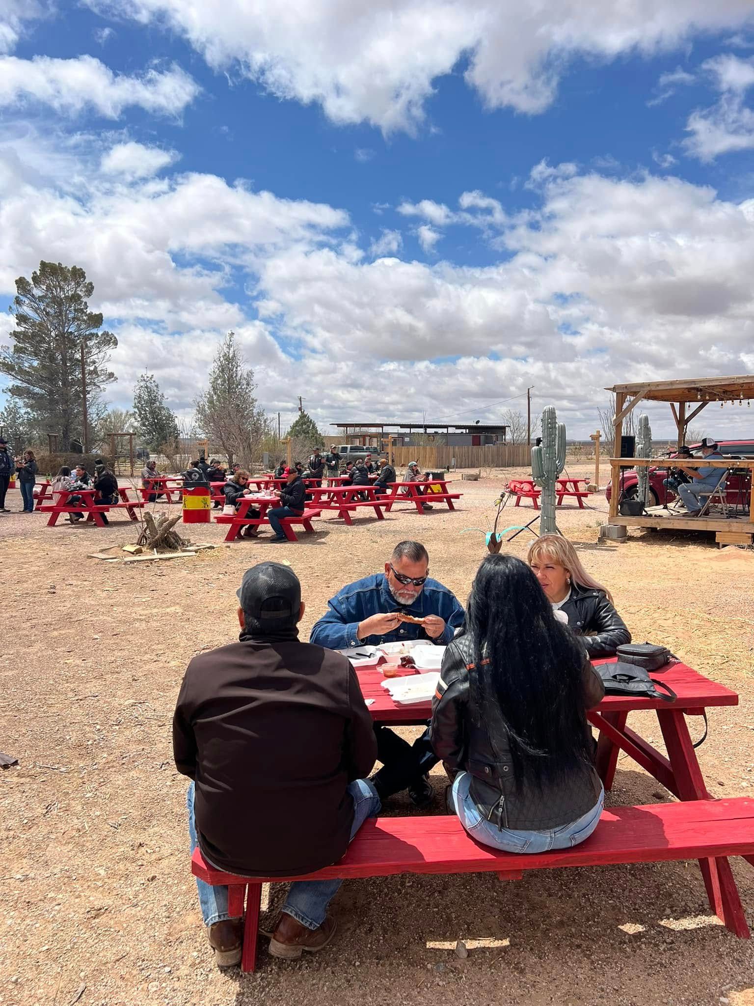 People seated at red picnic tables in a gravel yard under a cloudy blue sky, with equipment in the background.