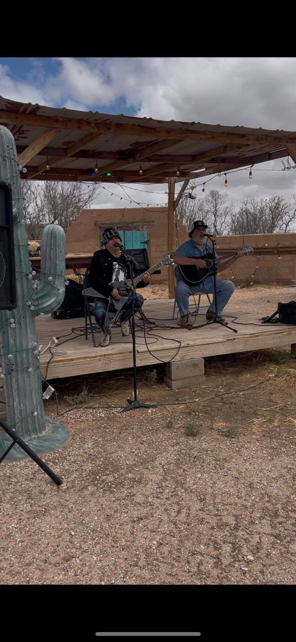 Two people seated on a rustic outdoor stage under a wooden shelter, playing instruments in a desert setting.
