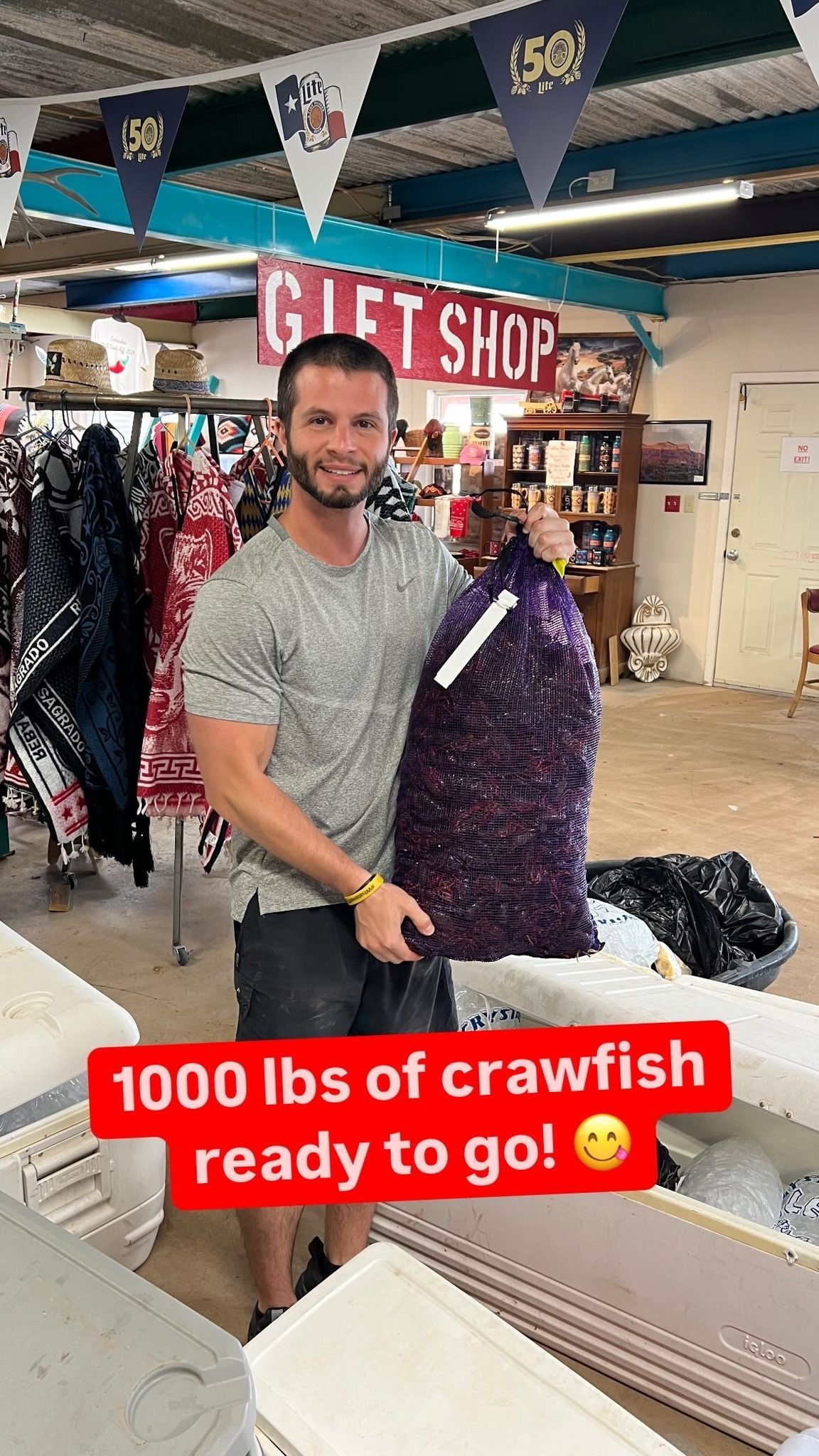 Man holding a purple crawfish sack in a gift shop, with “1000 lbs of crawfish ready to go!” text.