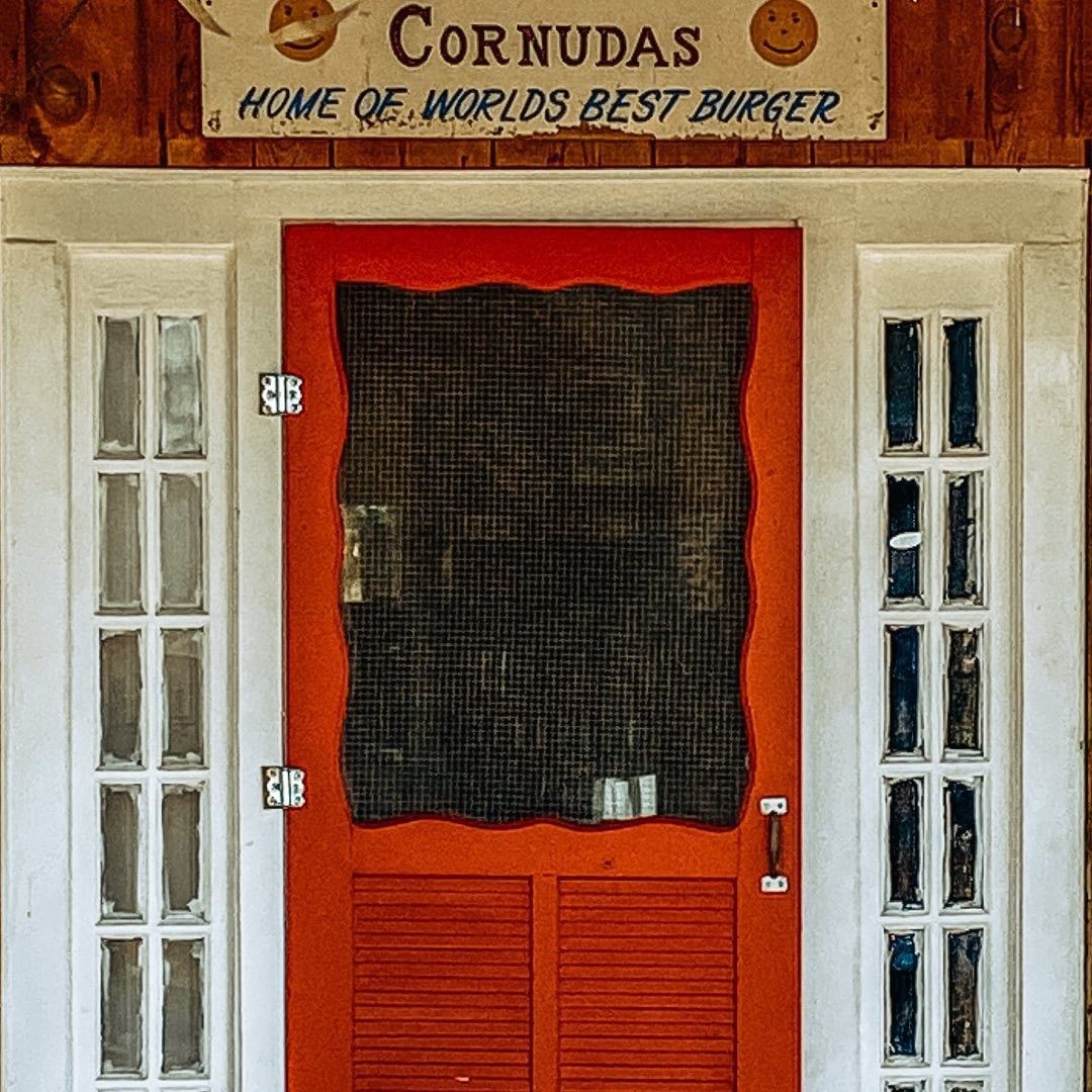 Red door with screen, flanked by white-framed windows beneath a sign reading “Cornudas” and “Home of world’s best burger.”