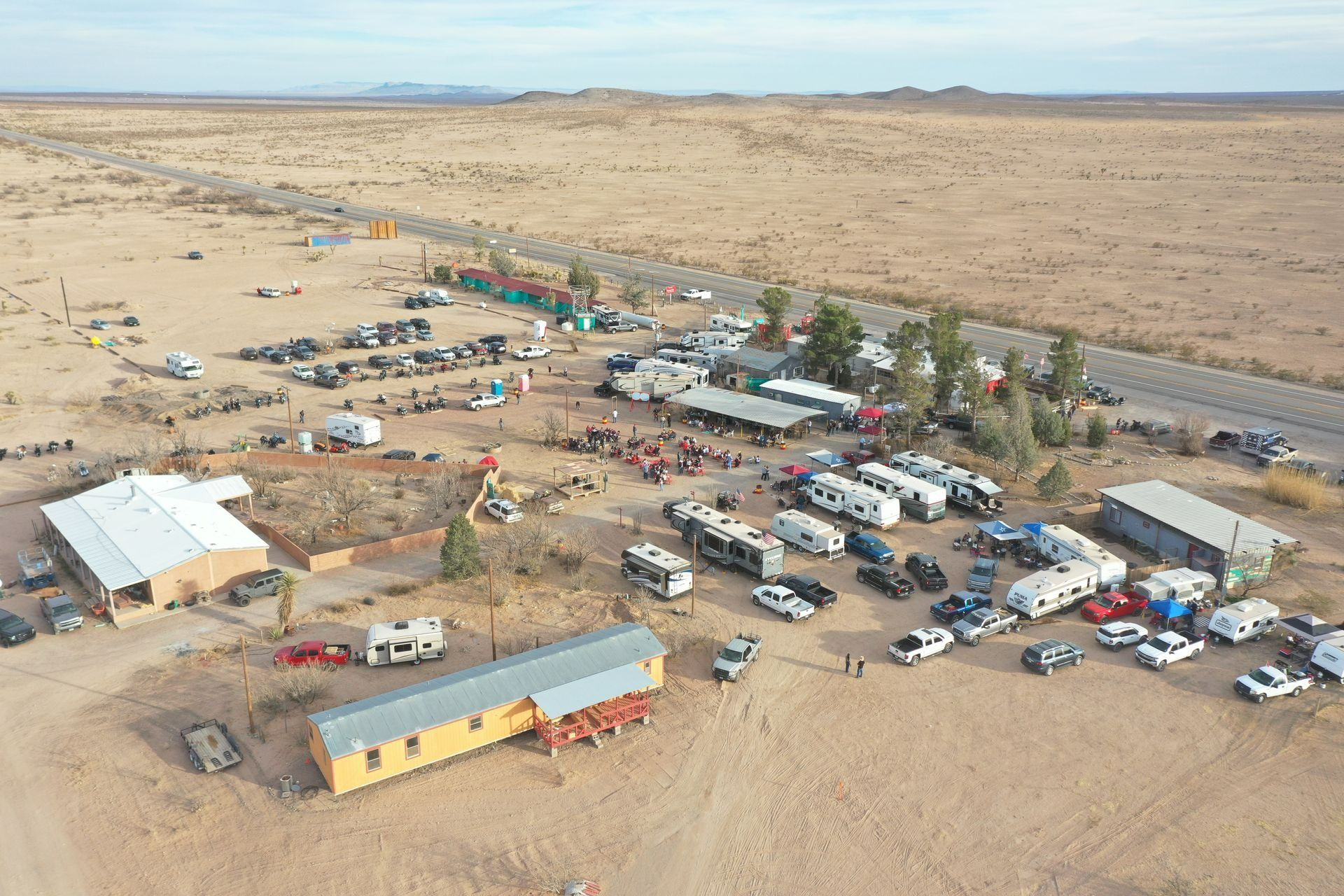 Aerial view of a desert campsite with many vehicles, tents, and buildings near a long road.
