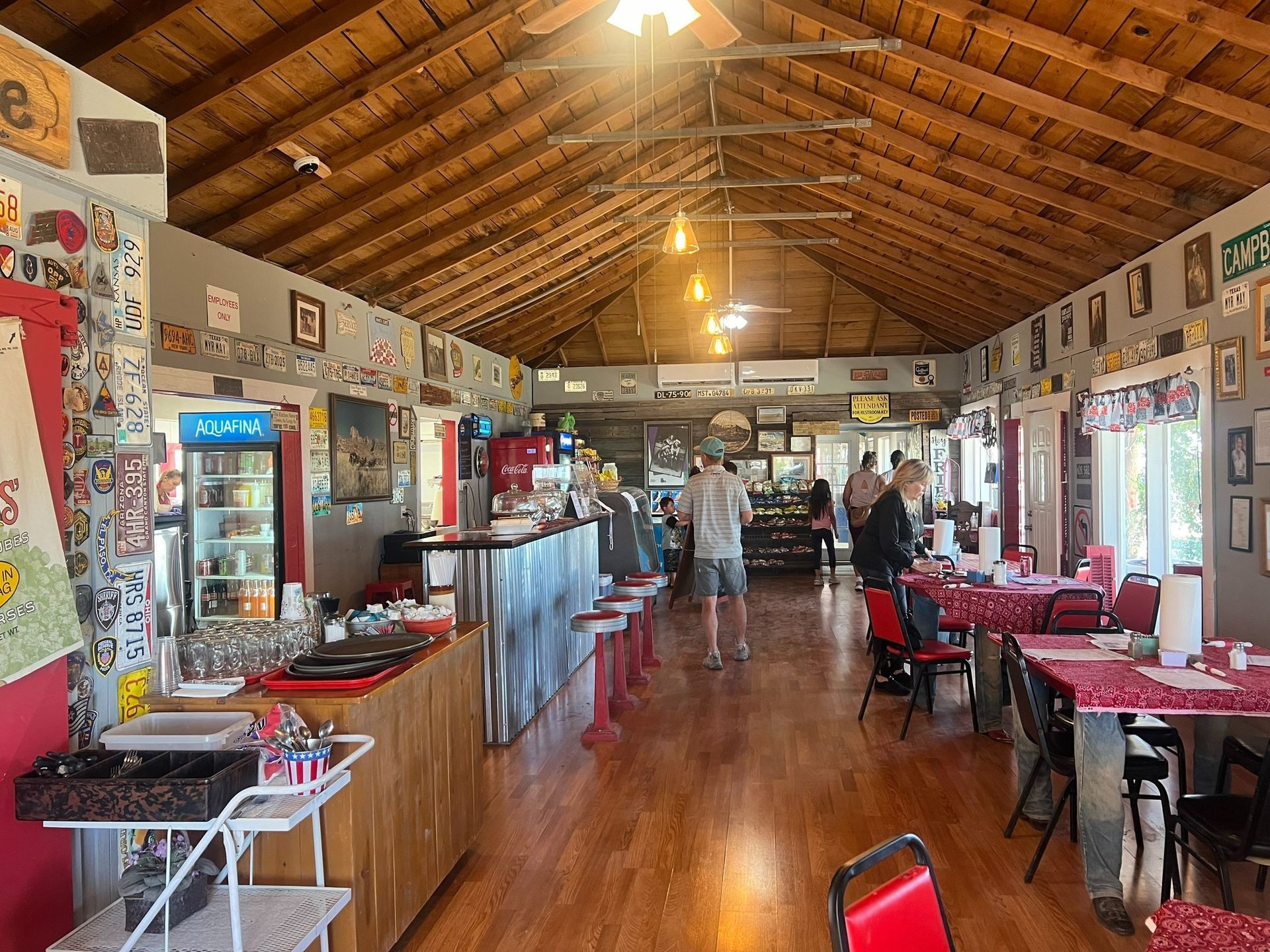 Long rustic dining hall with red chairs, wooden tables, and a high timber ceiling; a few people stand at the far end