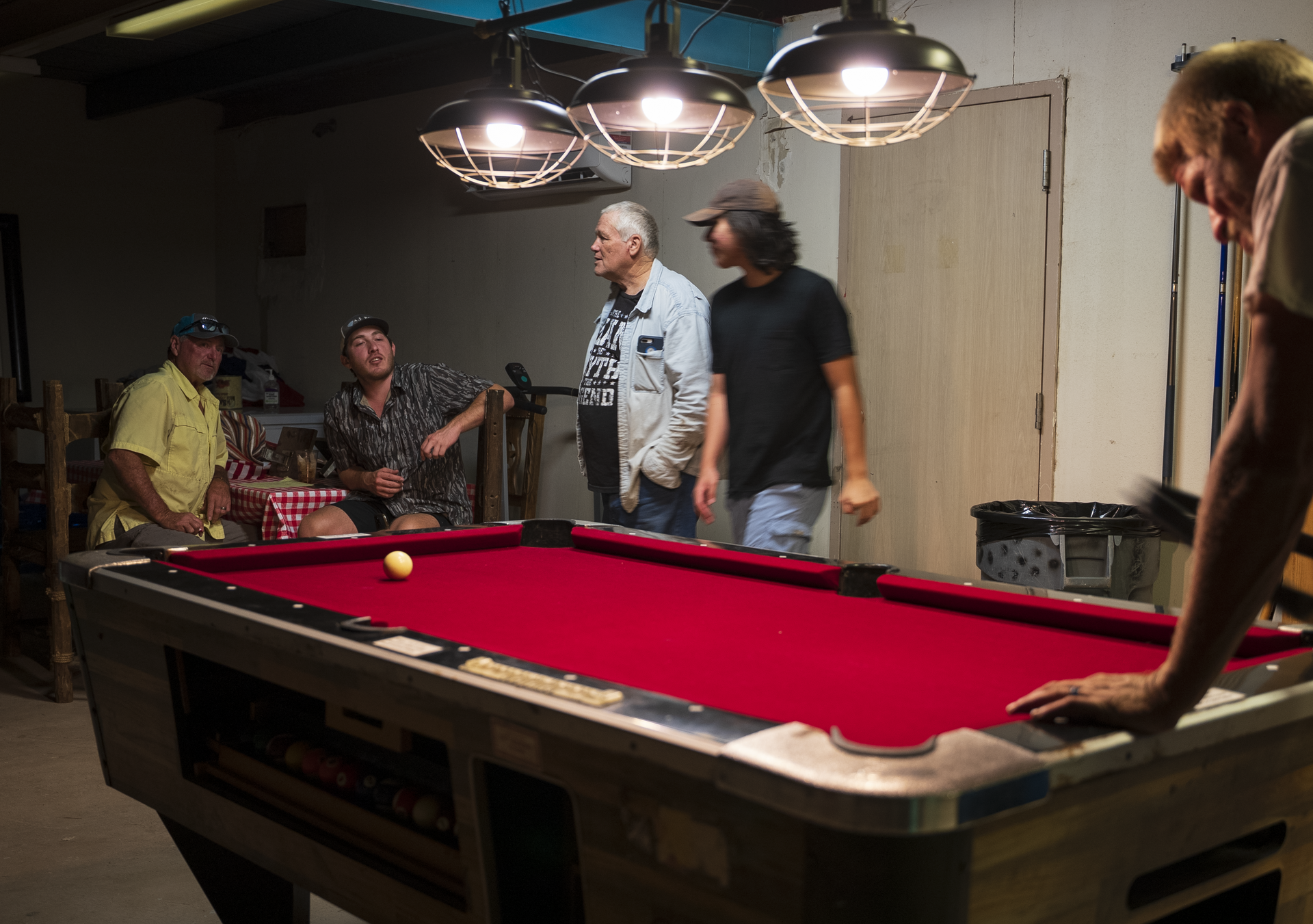 Men gathered around a red pool table in a dimly lit game room, with overhead lights shining down.