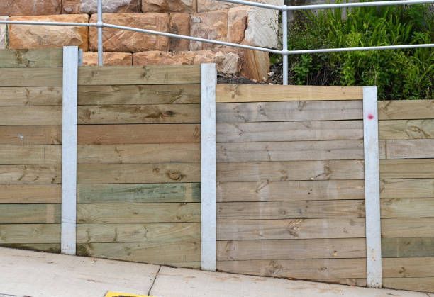Wooden retaining wall with metal supports, next to a stone wall, trees, and a sidewalk.