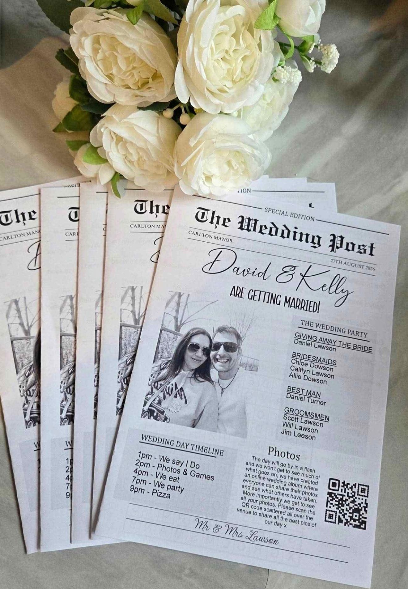 Wedding invitations with white roses and a newspaper-style layout on a light surface