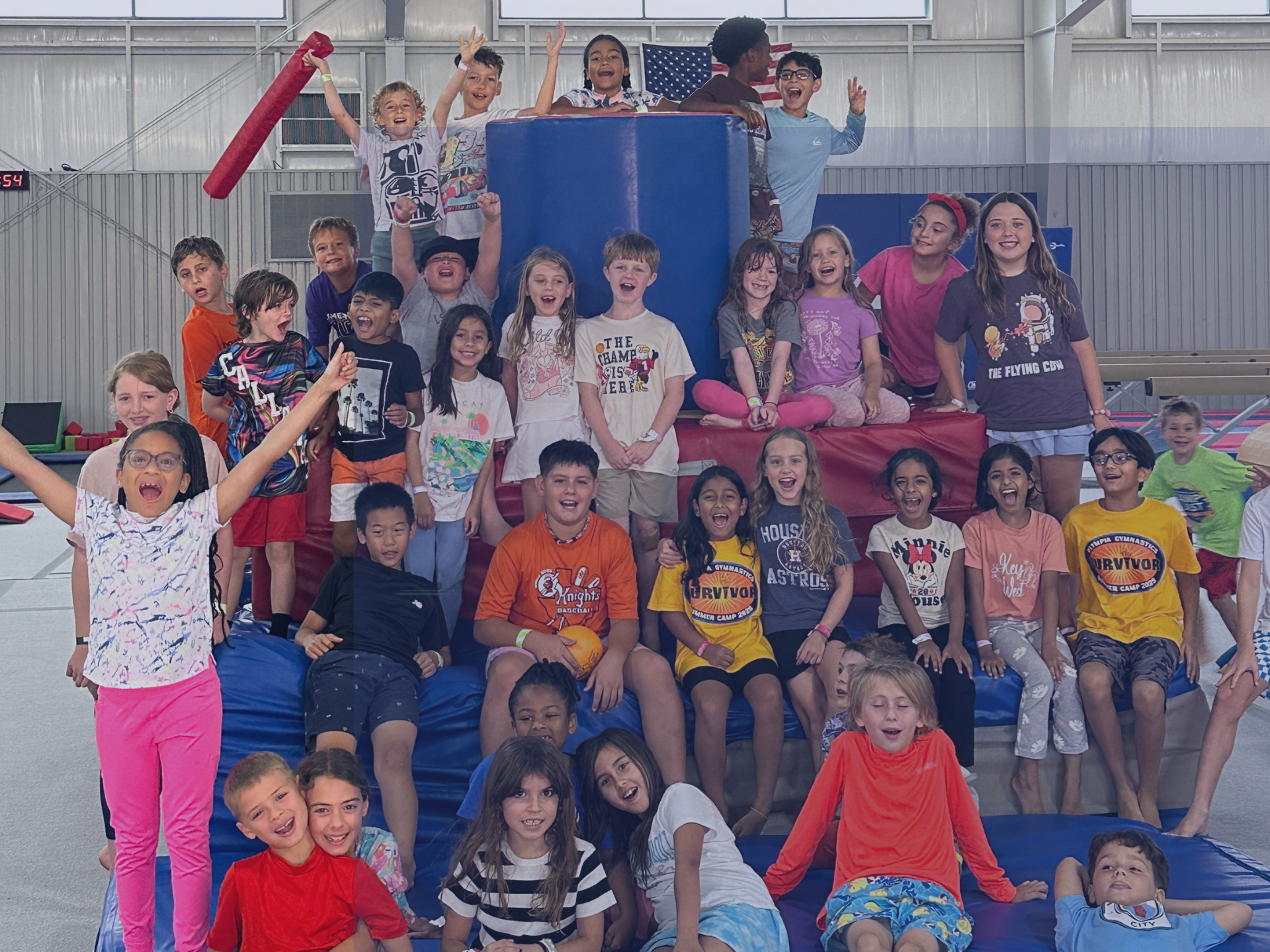 Group of kids at a gymnastics summer camp smiling and cheering while sitting and climbing on padded