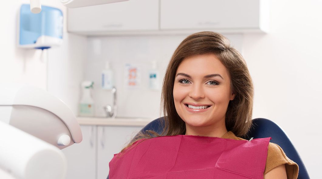 Paziente sorridente seduta sulla poltrona del dentista, con indosso un bavaglino rosa, e uno studio dentistico sullo sfondo.