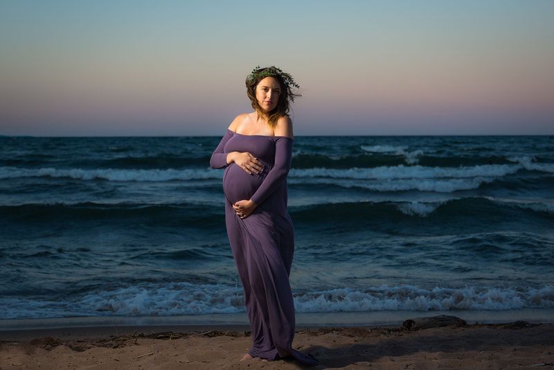 A pregnant woman in a purple dress is standing on the beach holding her belly.
