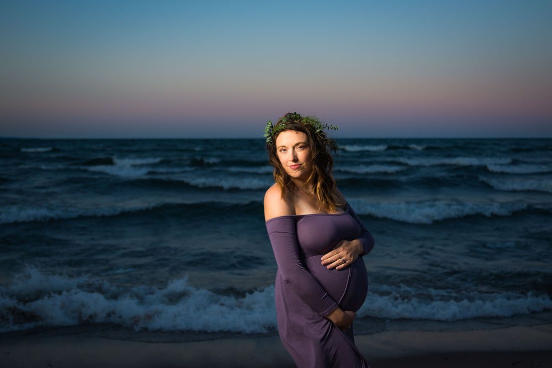 A pregnant woman in a purple dress is standing on the beach holding her belly.