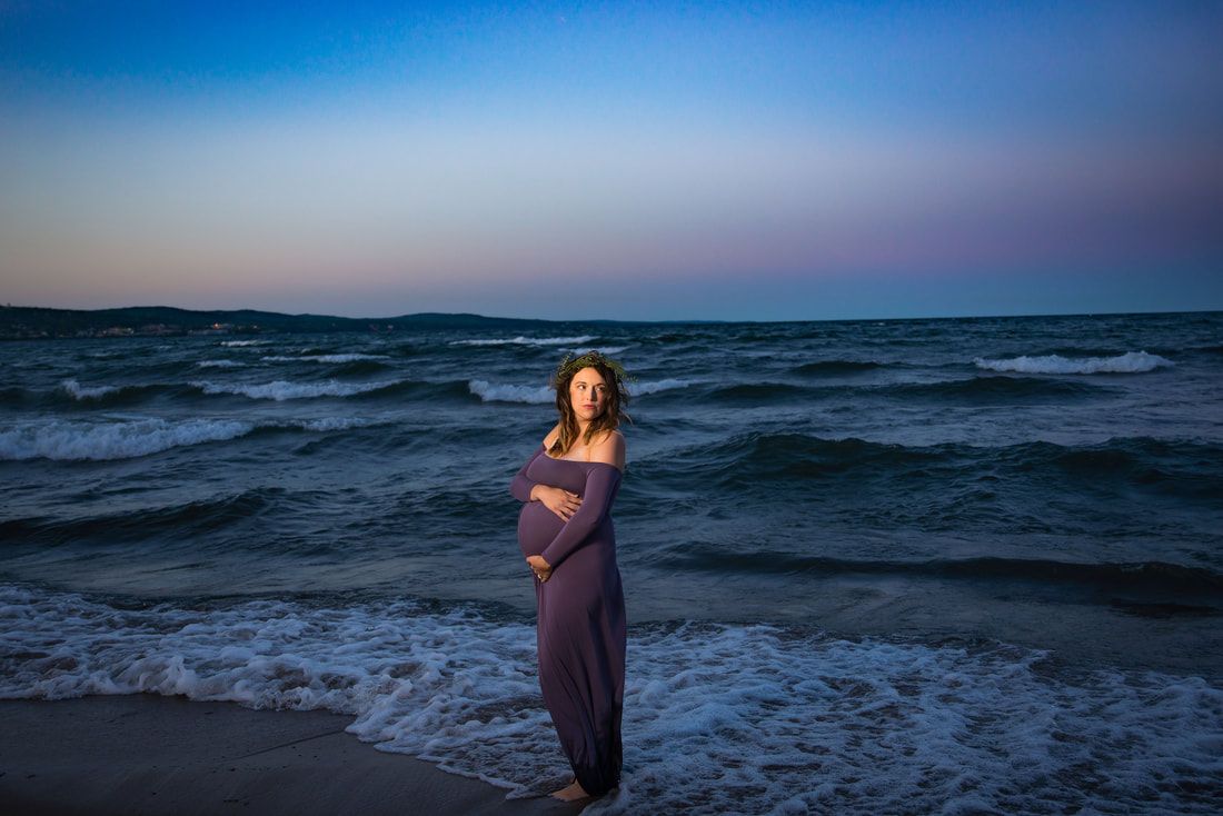 A pregnant woman in a purple dress is standing on the beach holding her belly.