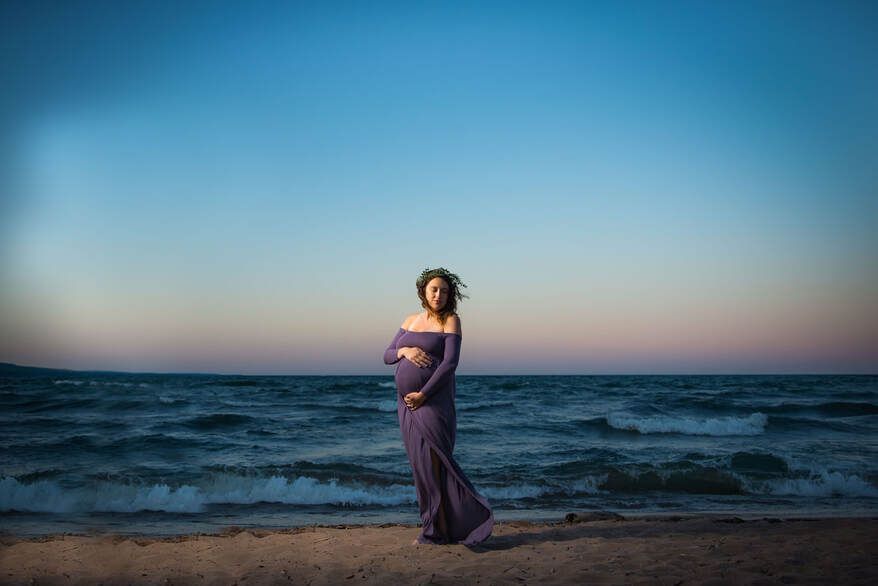 A pregnant woman in a purple dress is standing on the beach holding her belly.