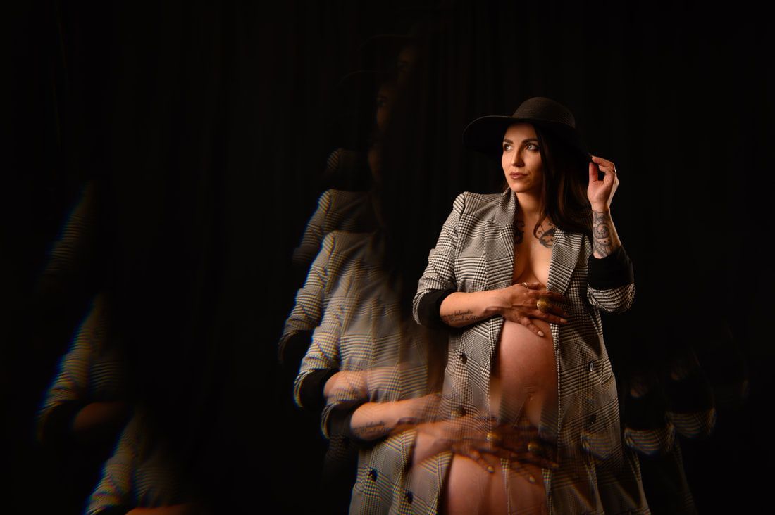 A pregnant woman wearing a hat and a trench coat is standing in a dark room.