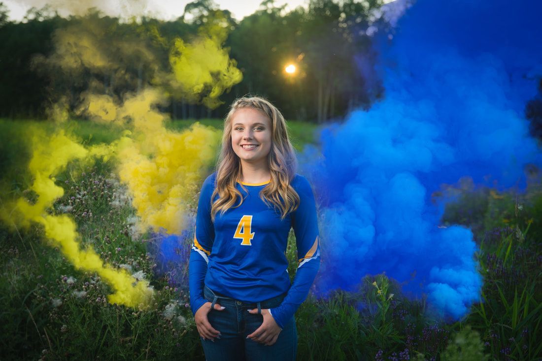 A girl is standing in a field with blue and yellow smoke in the background.