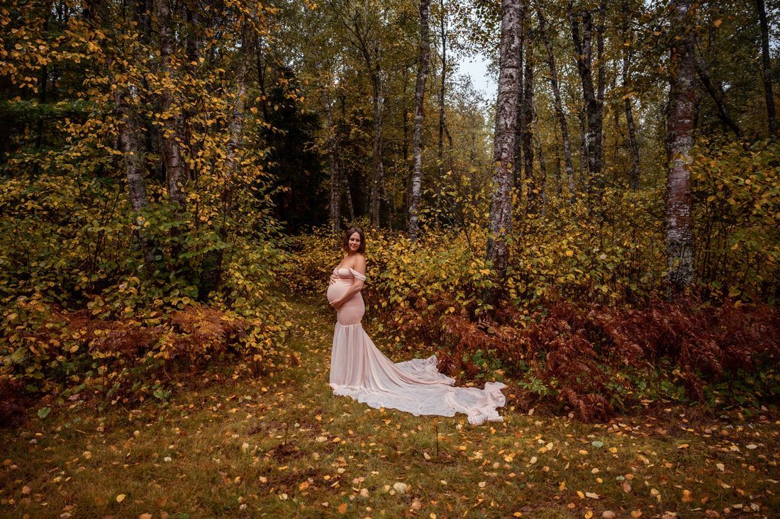 A pregnant woman in a long white dress is standing in the woods.