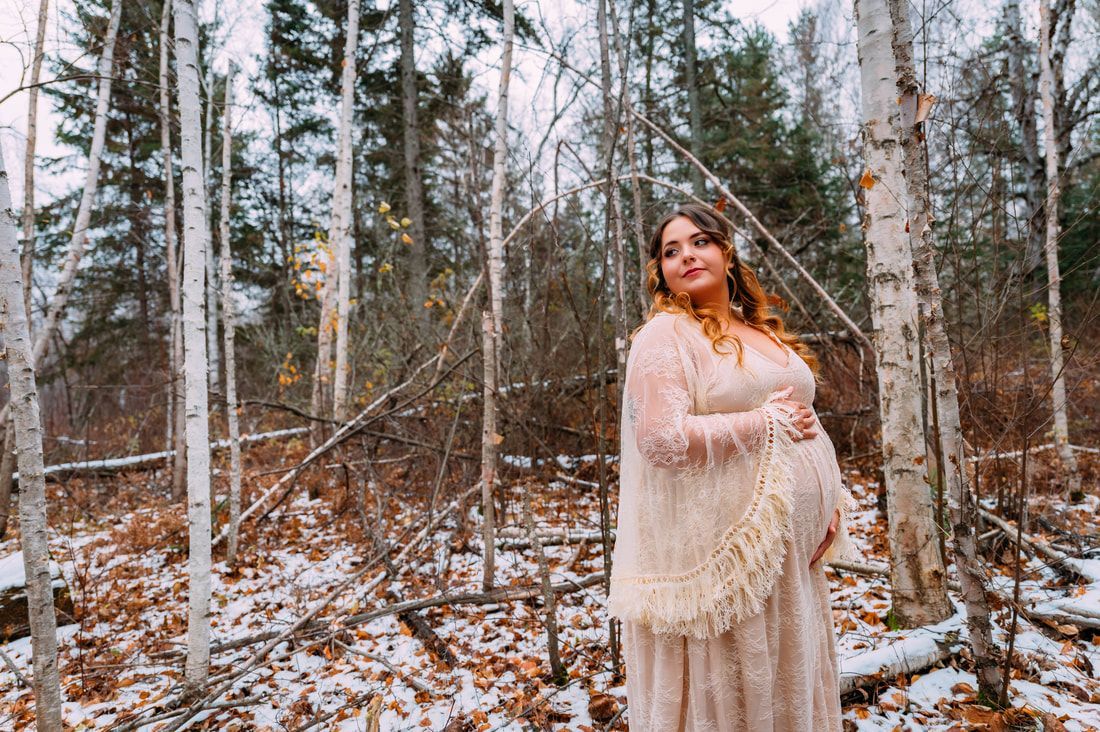 A pregnant woman in a white dress is standing in the woods holding her belly.