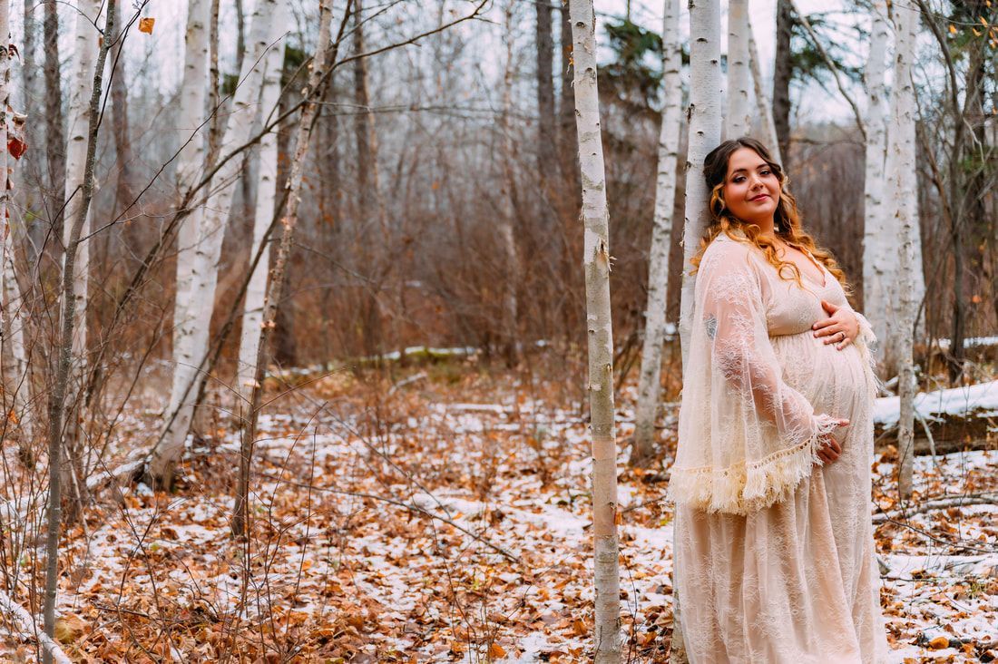 A pregnant woman in a white dress is standing in a snowy forest.