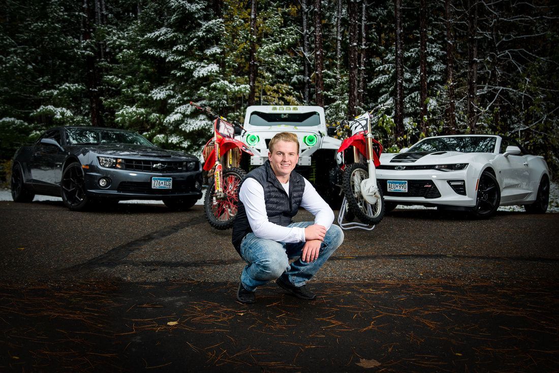 A man is squatting in front of three cars and a motorcycle.