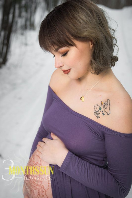 A pregnant woman in a purple dress is standing in the snow holding her belly.