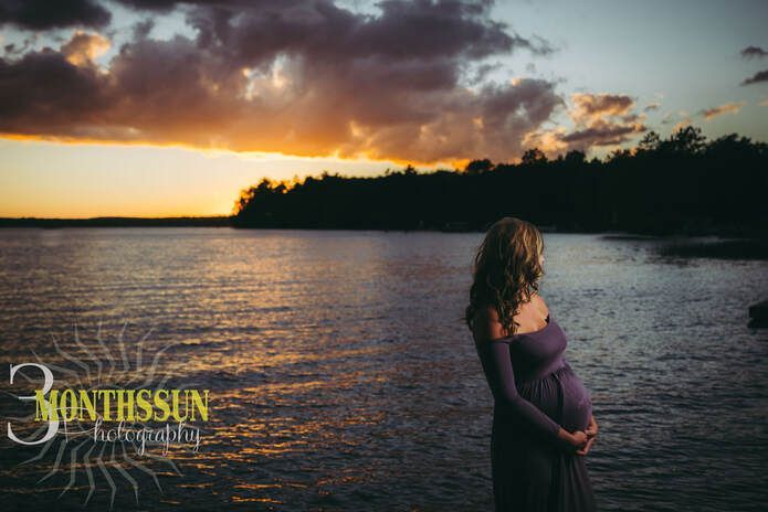 A pregnant woman in a purple dress is standing on the shore of a lake at sunset.