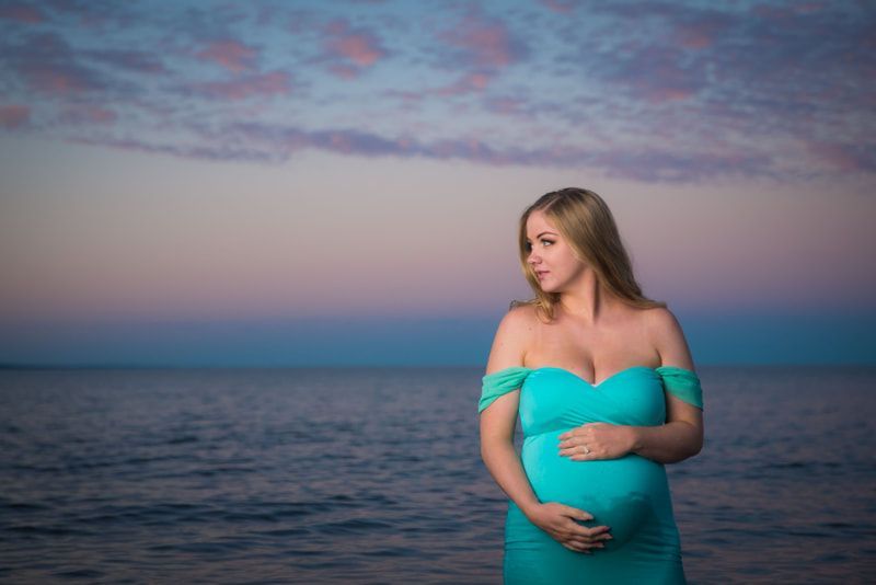 A pregnant woman in a blue dress is standing on the beach holding her belly.