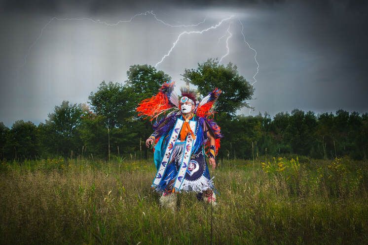 A man in a native american dress is standing in a field with lightning in the background.