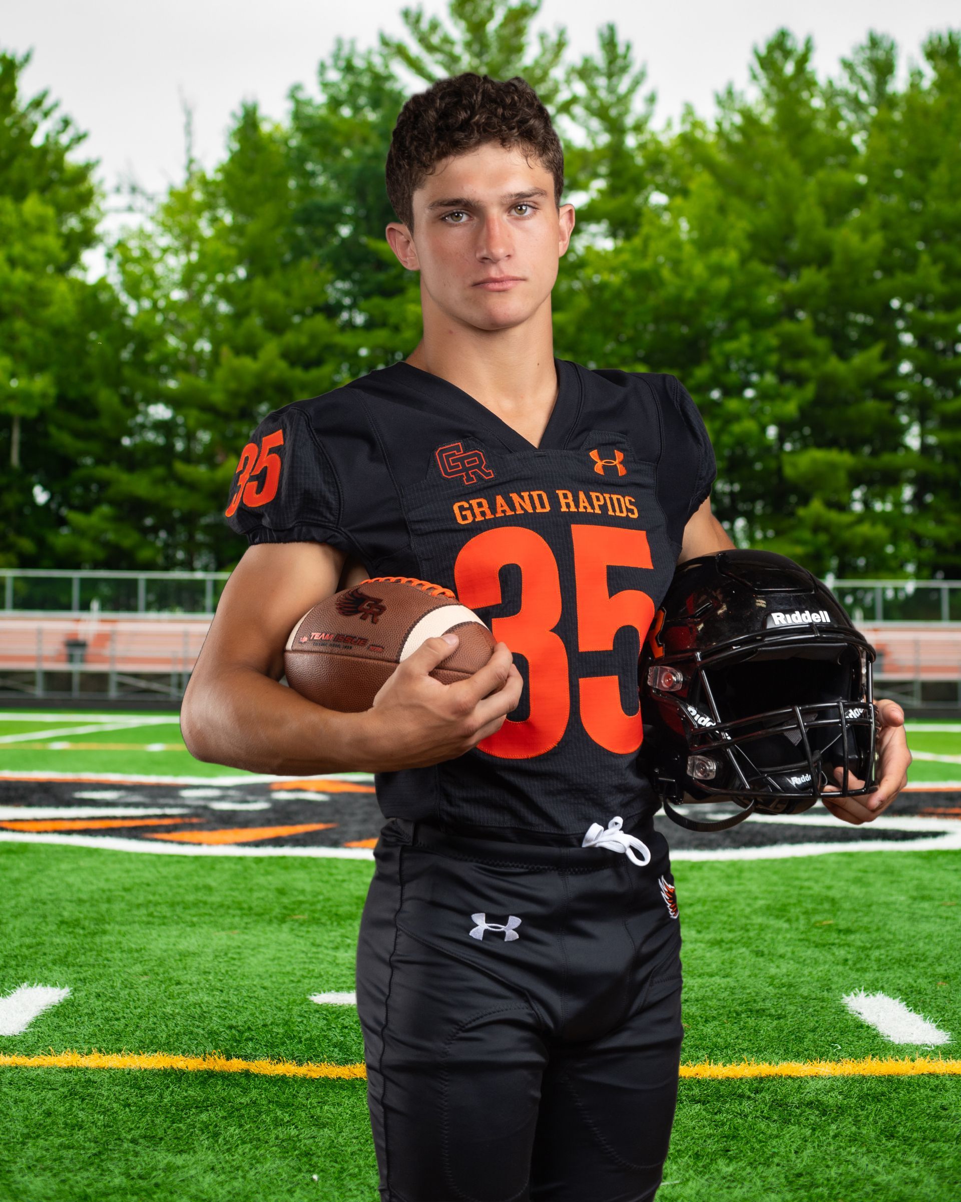 A young man in a football uniform is holding a football and a helmet on a field.