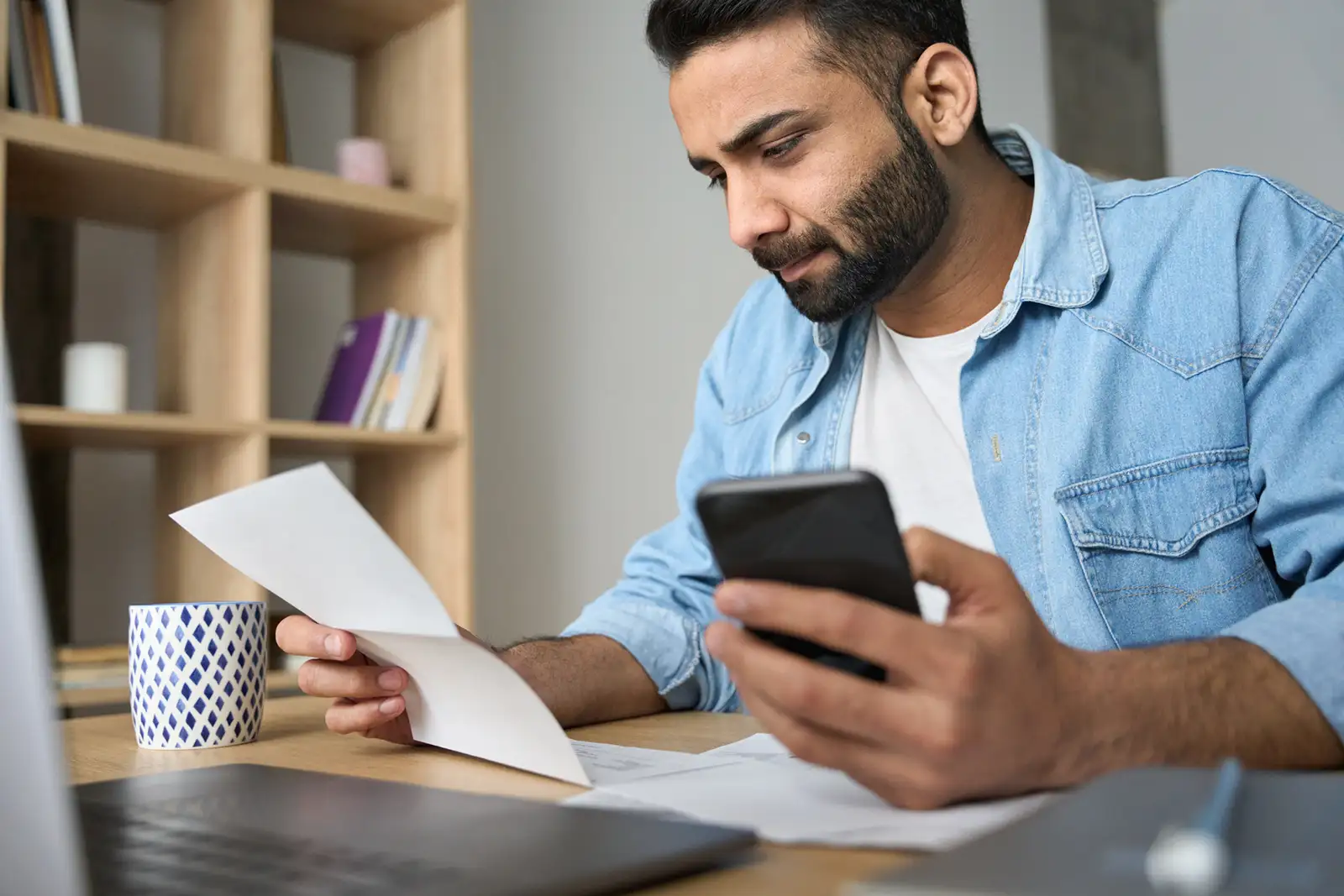 A man is sitting at a desk looking at a piece of paper and a cell phone.