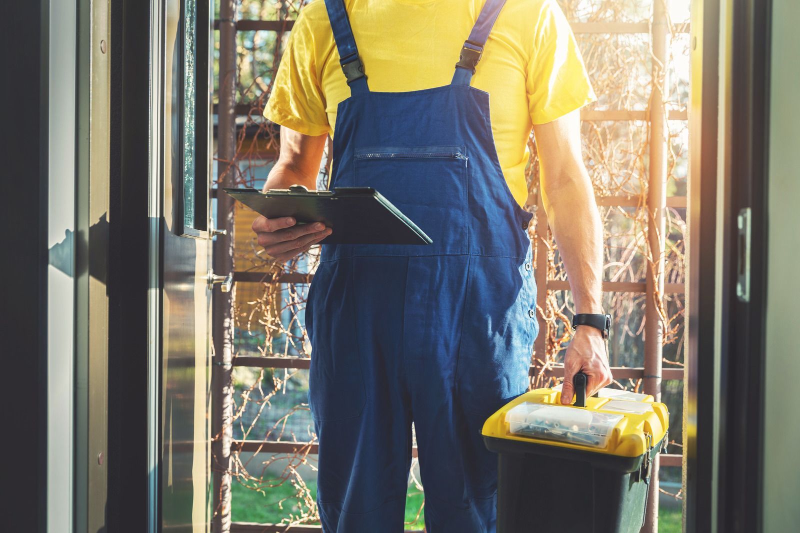 Delivery worker in blue overalls holding a tablet and package at a doorway
