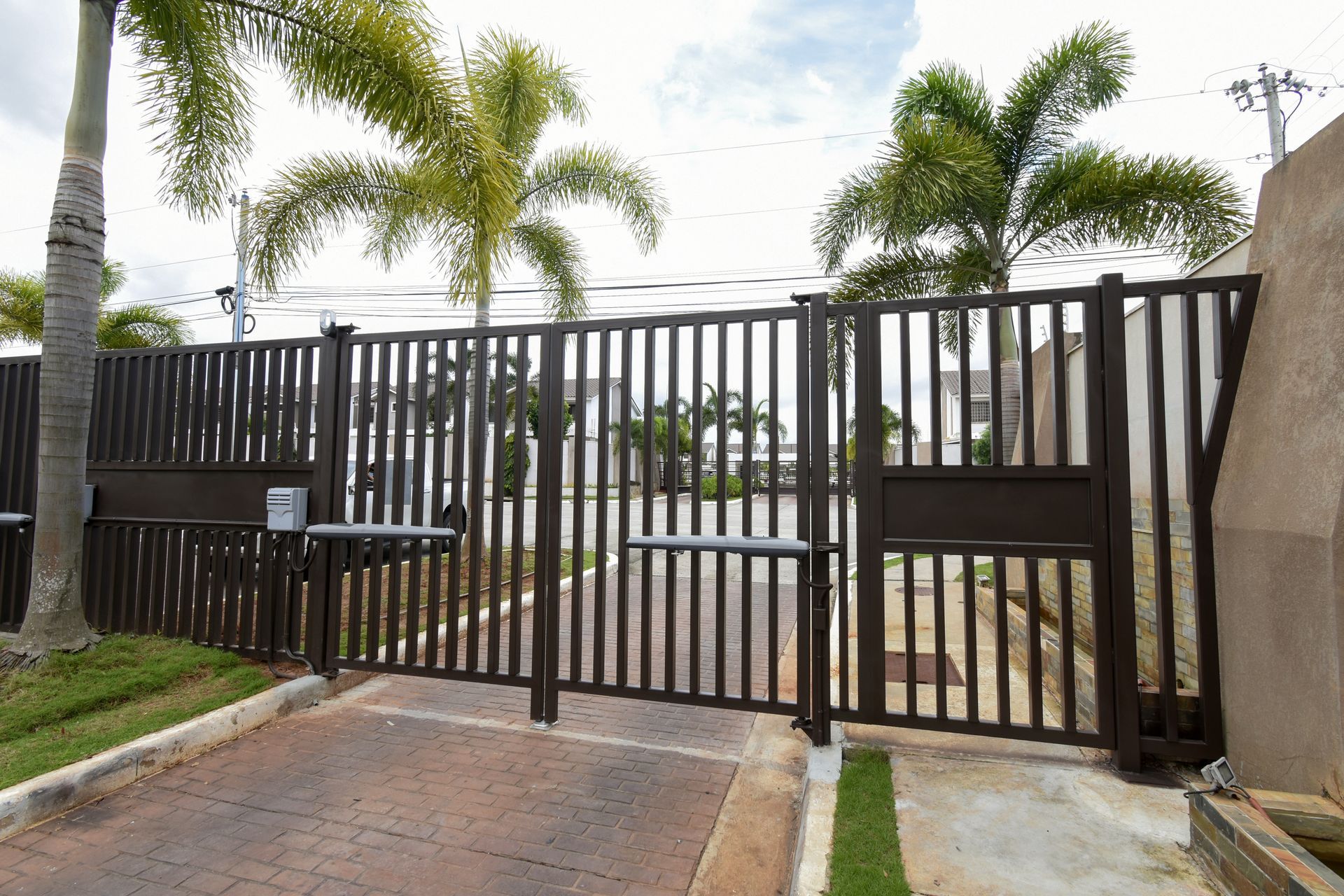 Black metal driveway gate beside a paved entrance, with palm trees and a modern house behind it