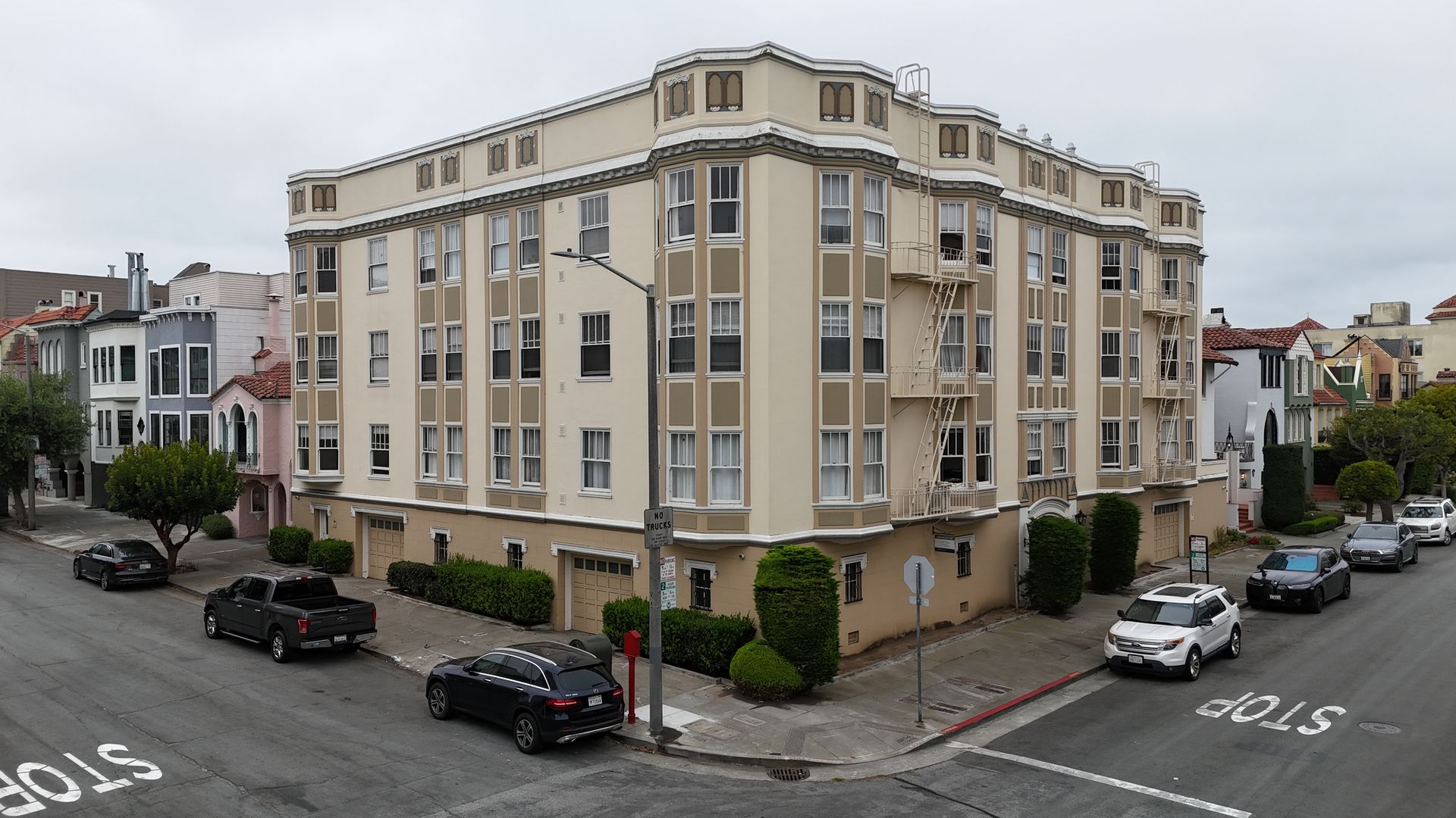 Corner beige apartment building on a city street with parked cars and overcast sky