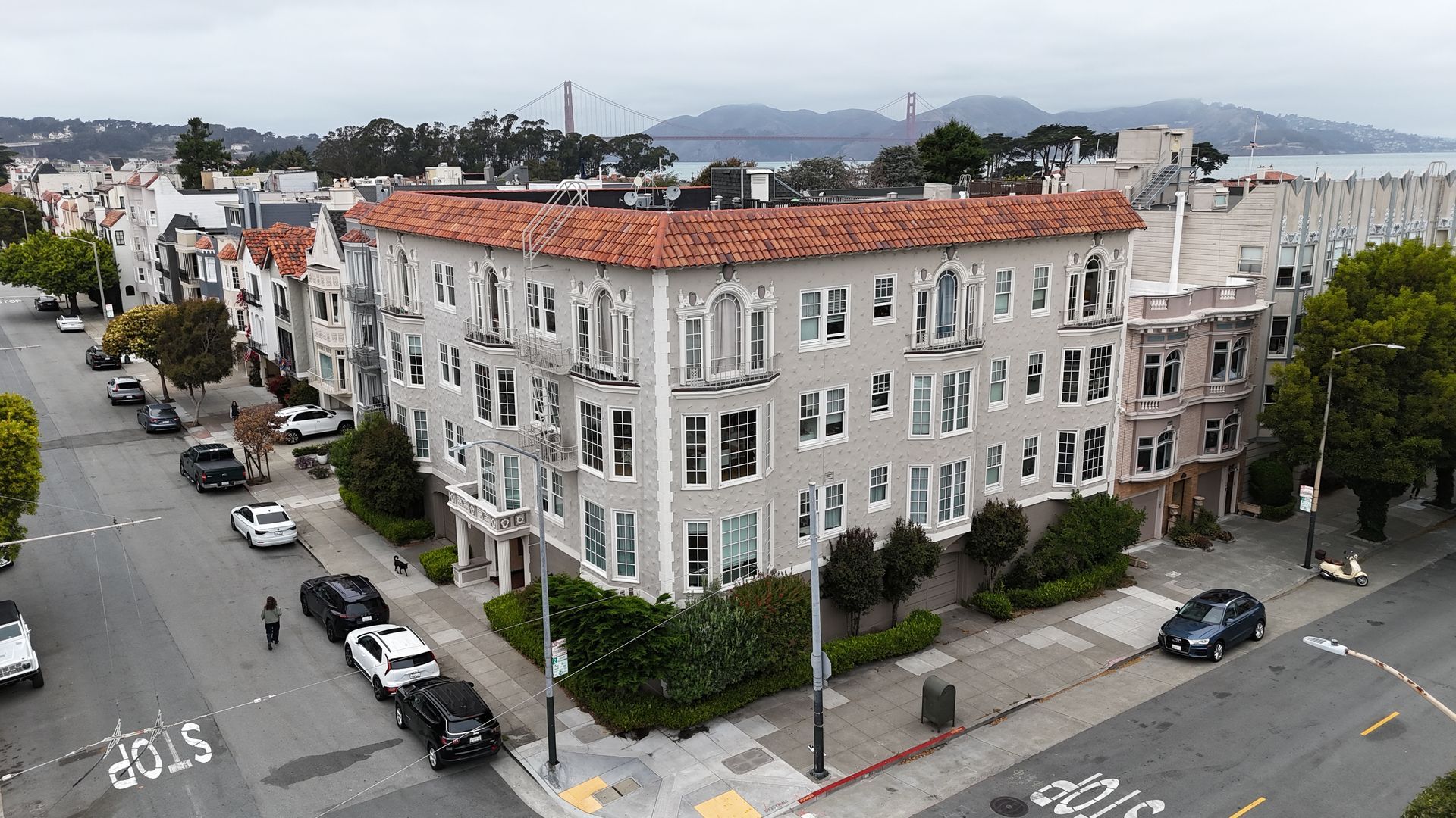 Corner view of a white apartment building with red tile roof on a city street, with parked cars and hills in back