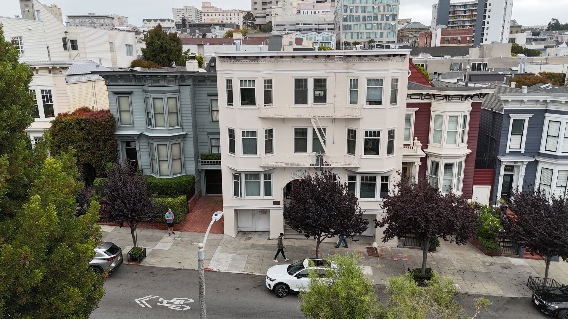 Apartment buildings lining a city street with parked cars and leafy trees under an overcast sky
