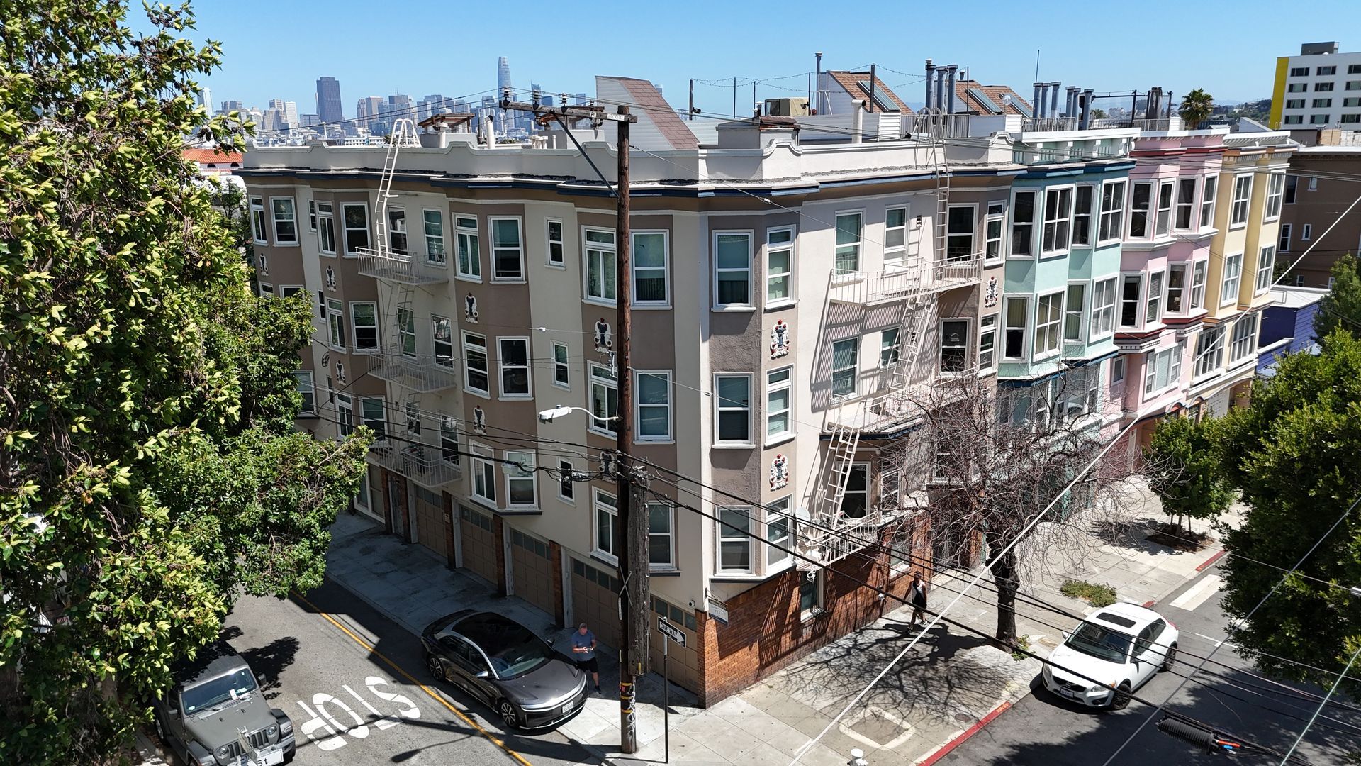 Corner apartment building on a sunny city street with parked cars and trees, skyline in the background