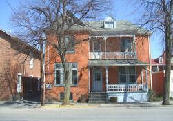 Brick two-story house with red shutters and a covered porch on a tree-lined street.