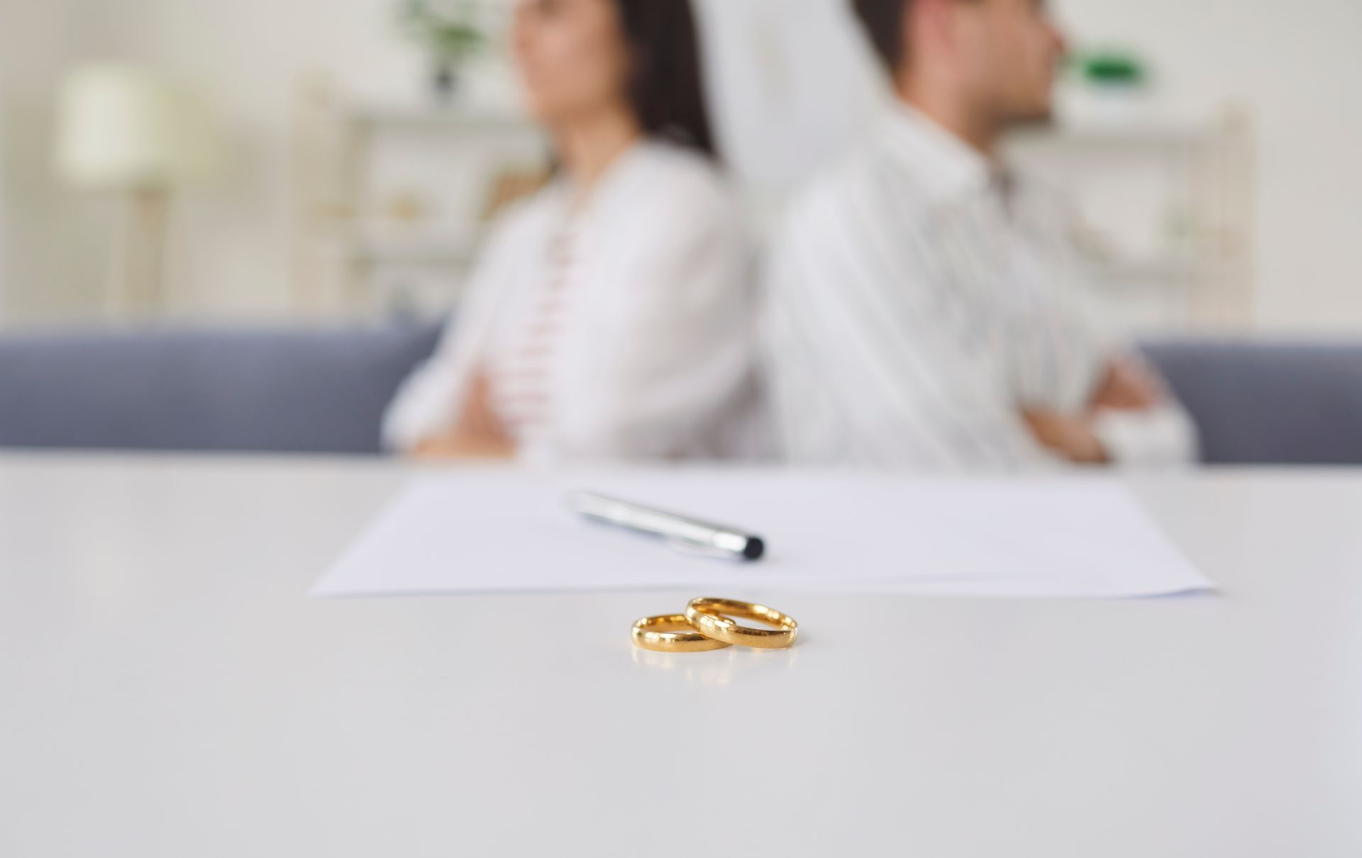 Gold wedding rings in the foreground, with a blurred couple sitting back-to-back in the background.