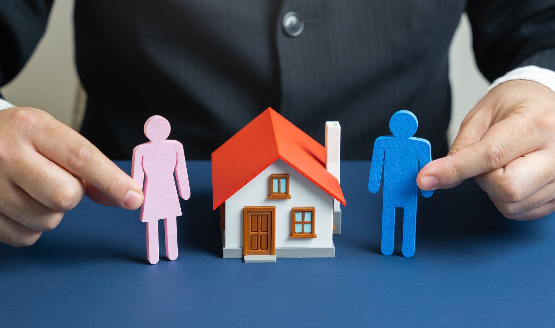 Hands holding pink and blue figures beside a small house model on a blue surface