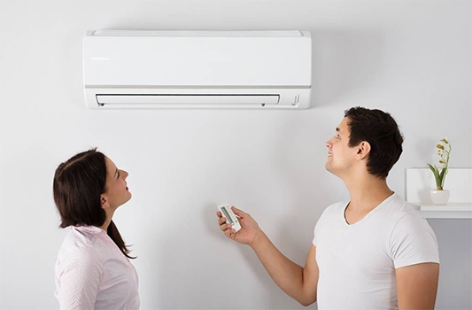 A Couple Looks Up at a Wall-mounted Air Conditioner as the Man Holds a Remote — Carl Voss Electrical In Urangan, QLD