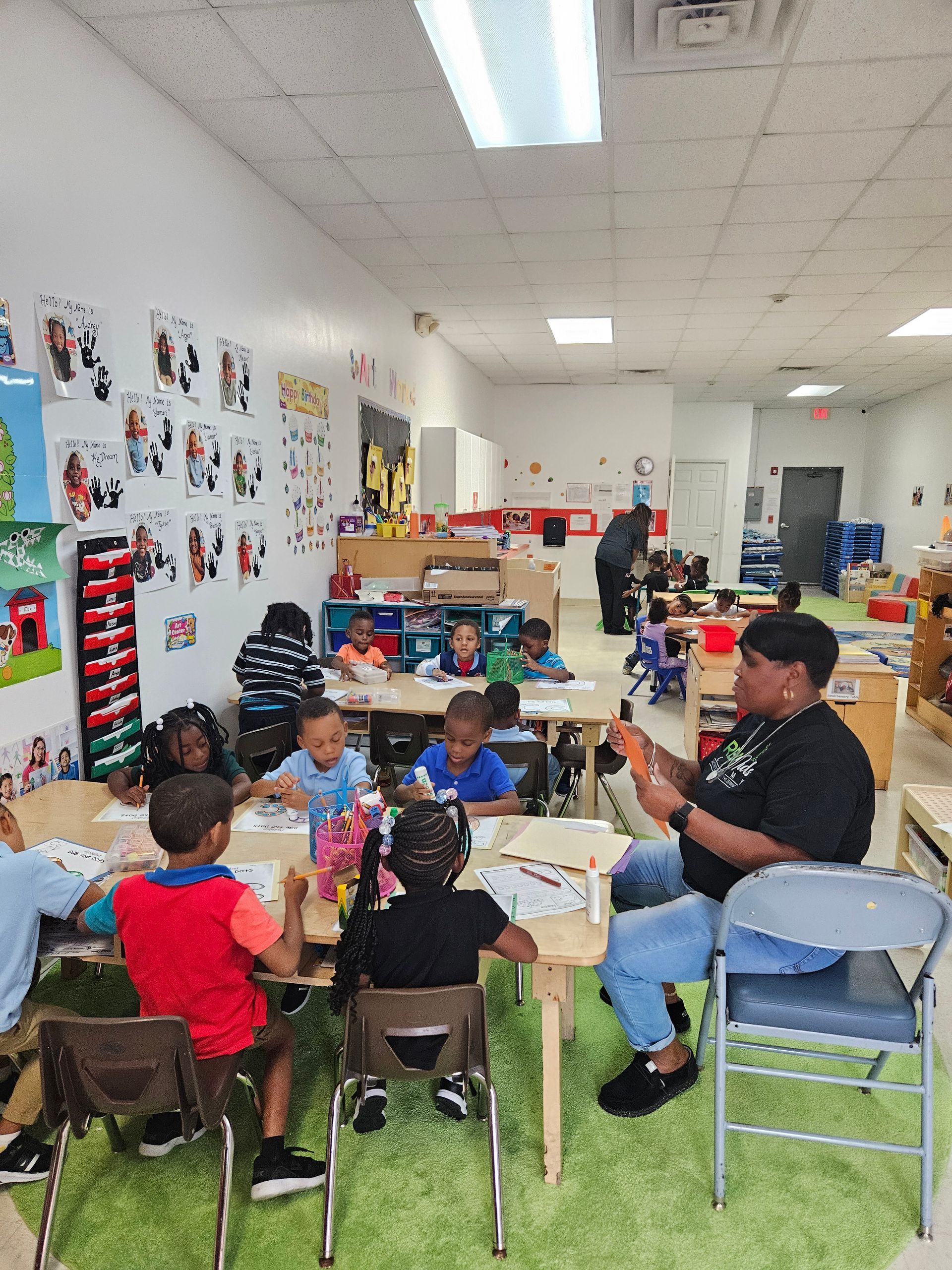 A group of children are sitting at tables in a classroom with a man sitting in a chair.