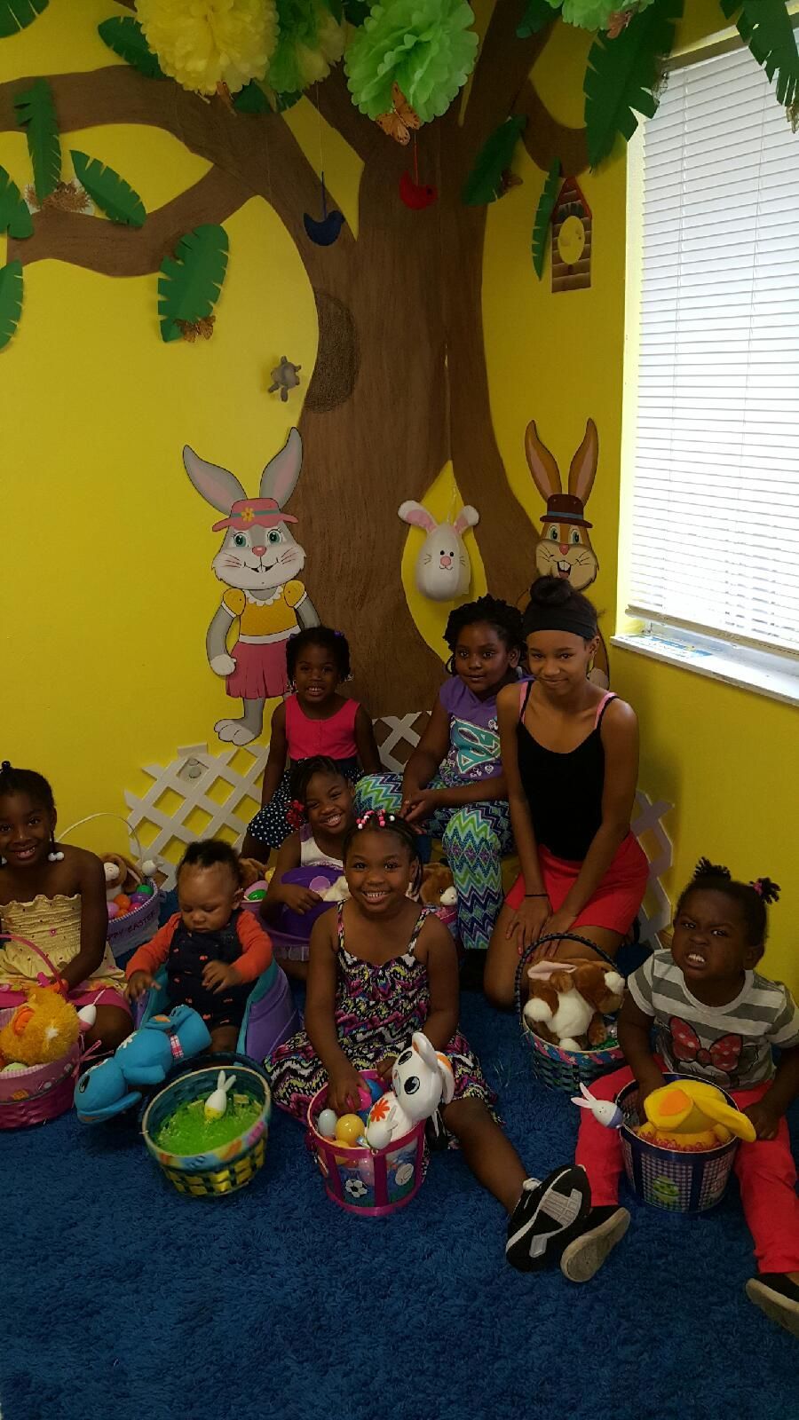 A group of children are sitting on the floor in a room with a tree on the wall.