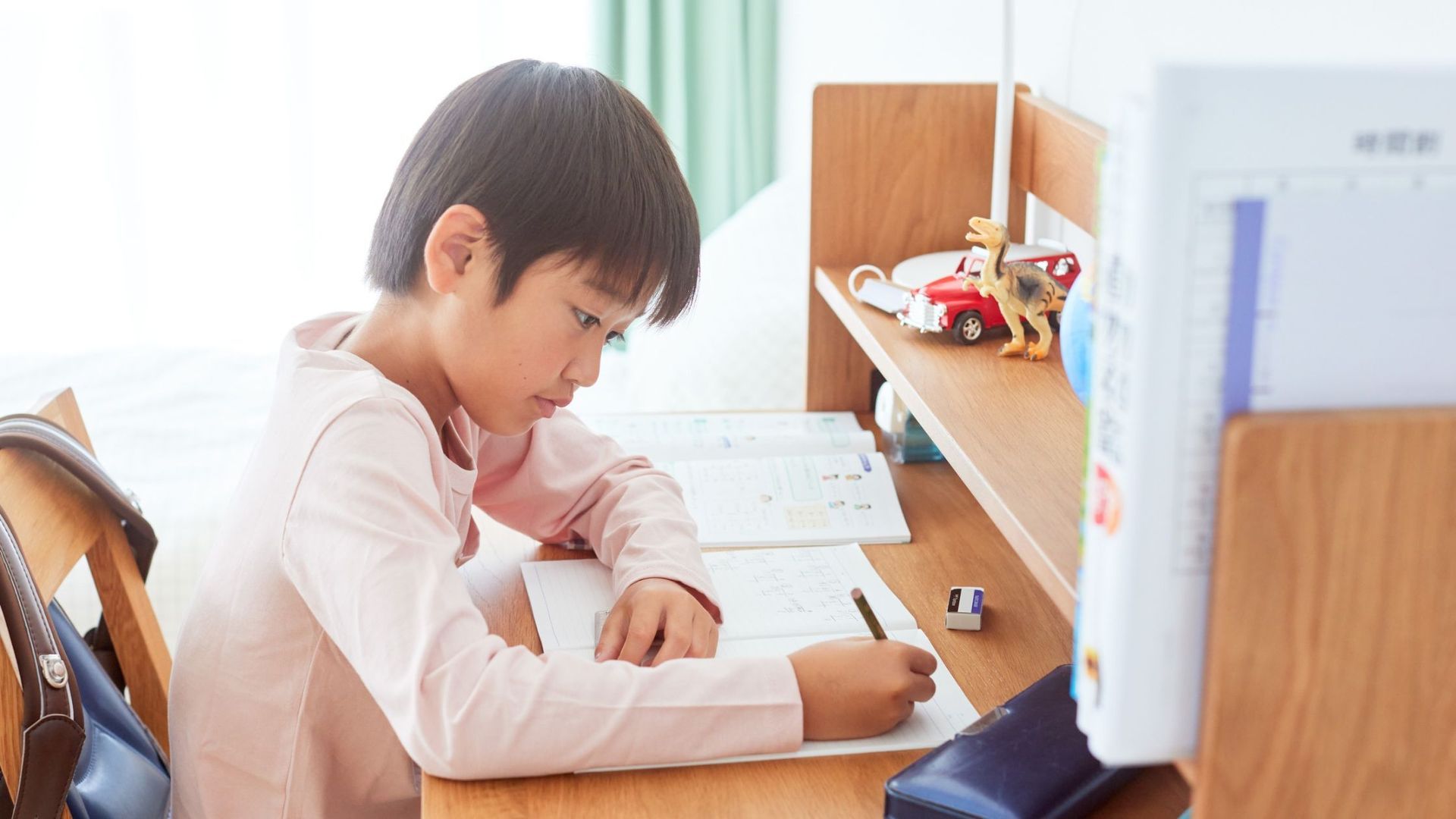 Boy writing at a wooden desk with books, toys, and a backpack in a room with natural light.
