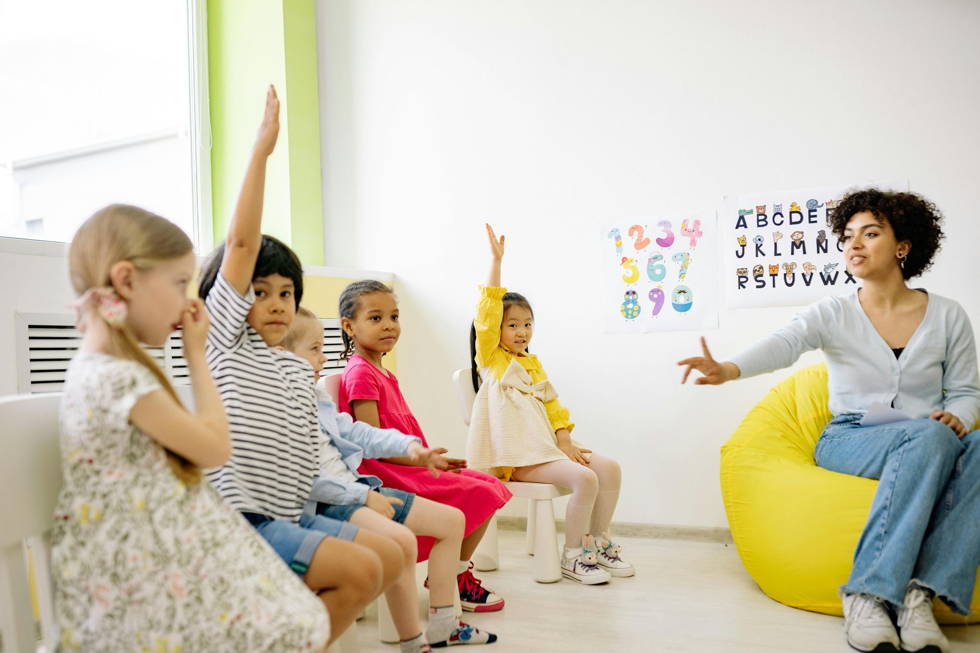 A group of children are sitting in chairs and raising their hands to answer a question.