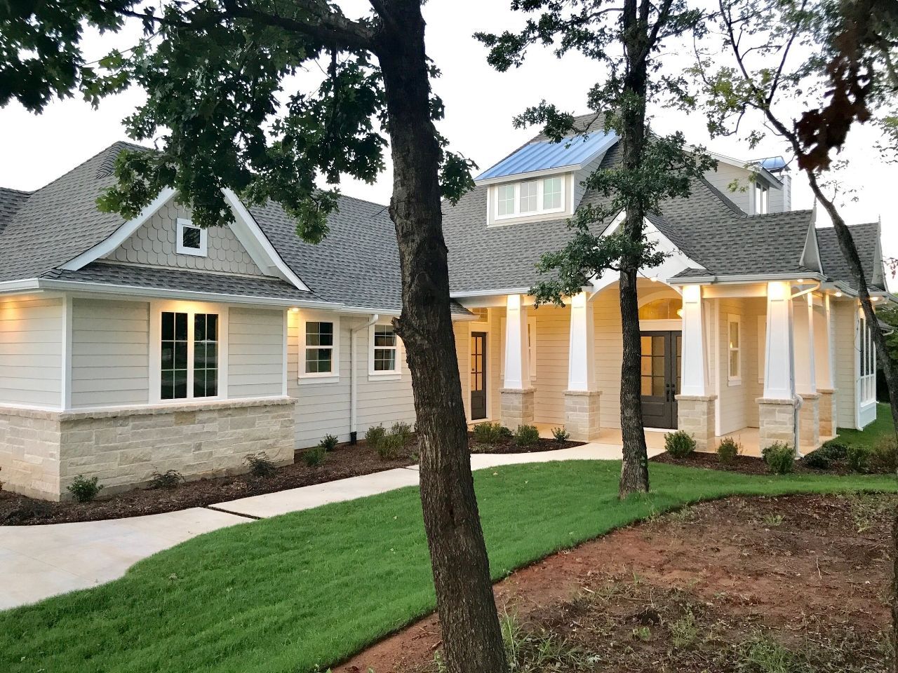 A light-colored house with a long driveway and green lawn surrounded by trees.