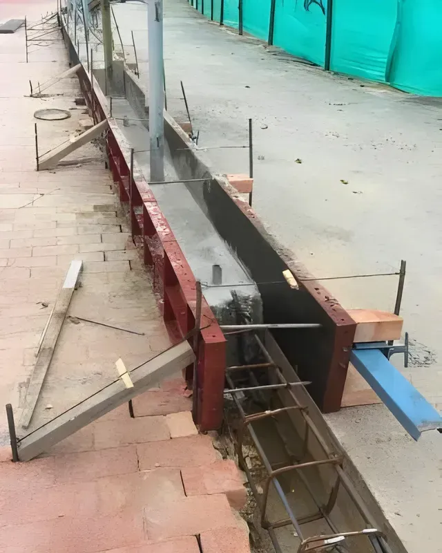 Acera dañada con barras de refuerzo expuestas y zanja de hormigón junto a un edificio en construcción.