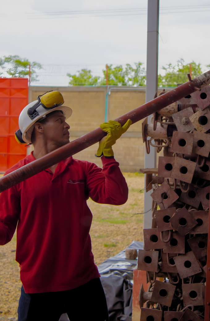 Un trabajador con camisa roja levanta un tubo metálico largo junto a una columna de ladrillos apilados en el exterior.
