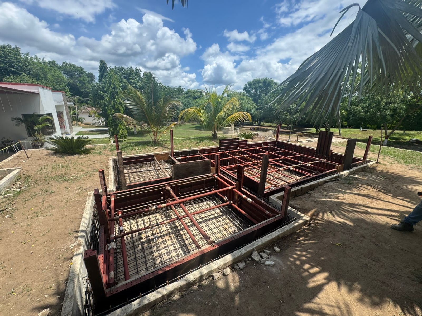 Cimentación de barras de refuerzo en construcción en un terreno soleado con palmeras y una casa parcialmente construida en las cercanías.