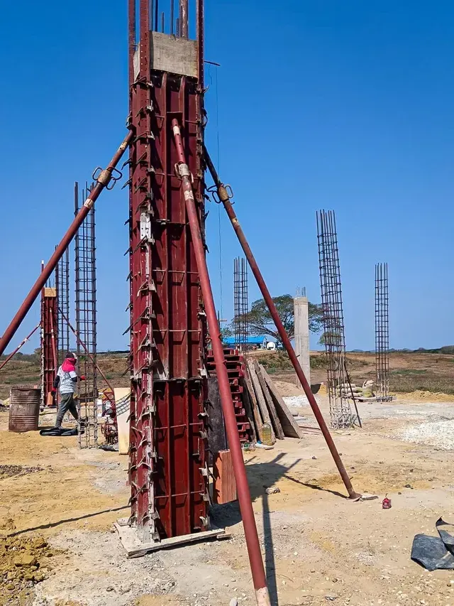 Obra en construcción con un pilote de cimentación de acero rojo y puntales de soporte bajo un cielo azul.