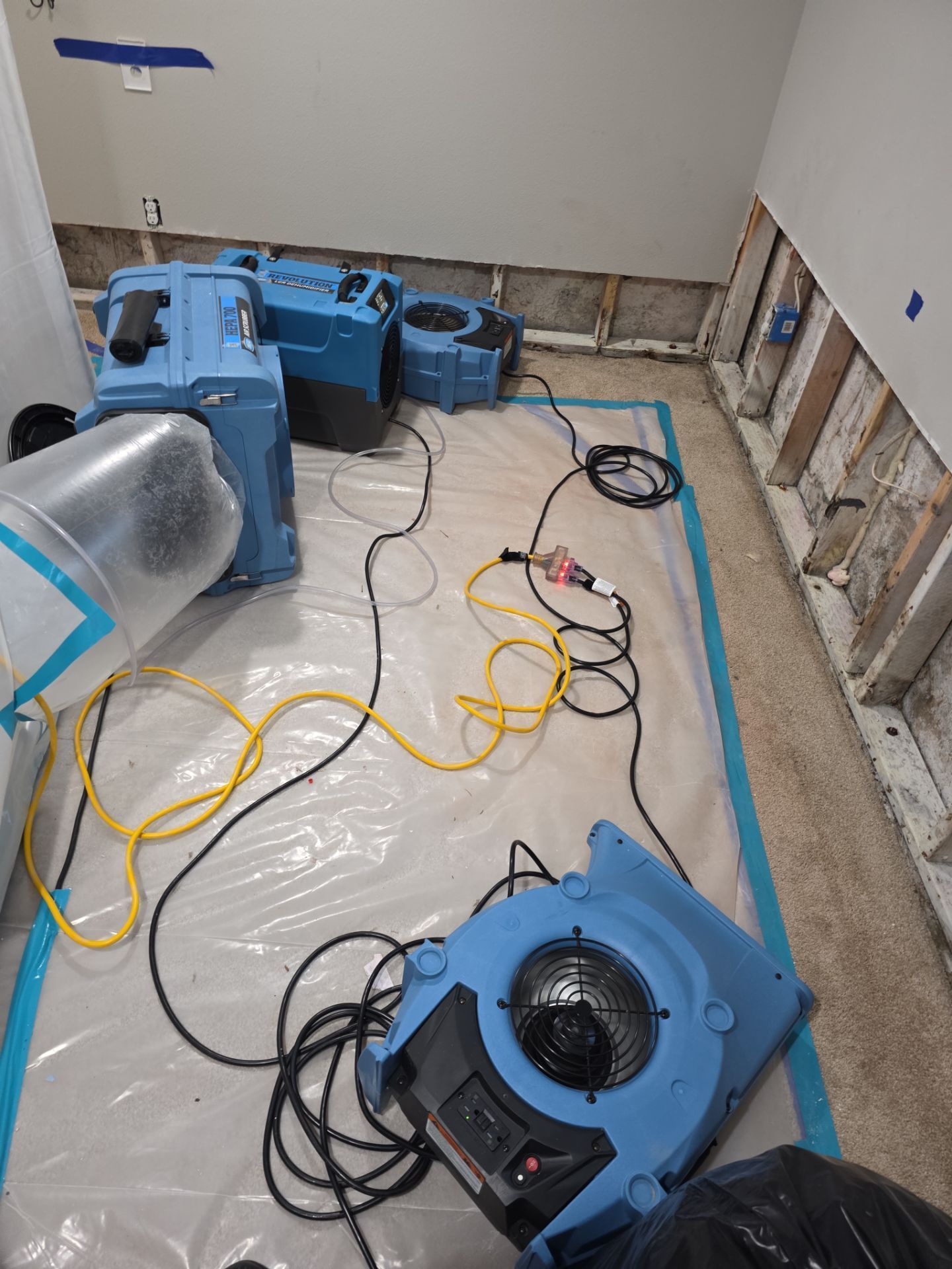 Several blue industrial fans and hoses on a floor covered with plastic sheeting in a room under renovation